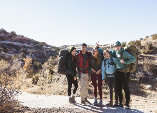 five young men and women with backpacks standing in a desert scene